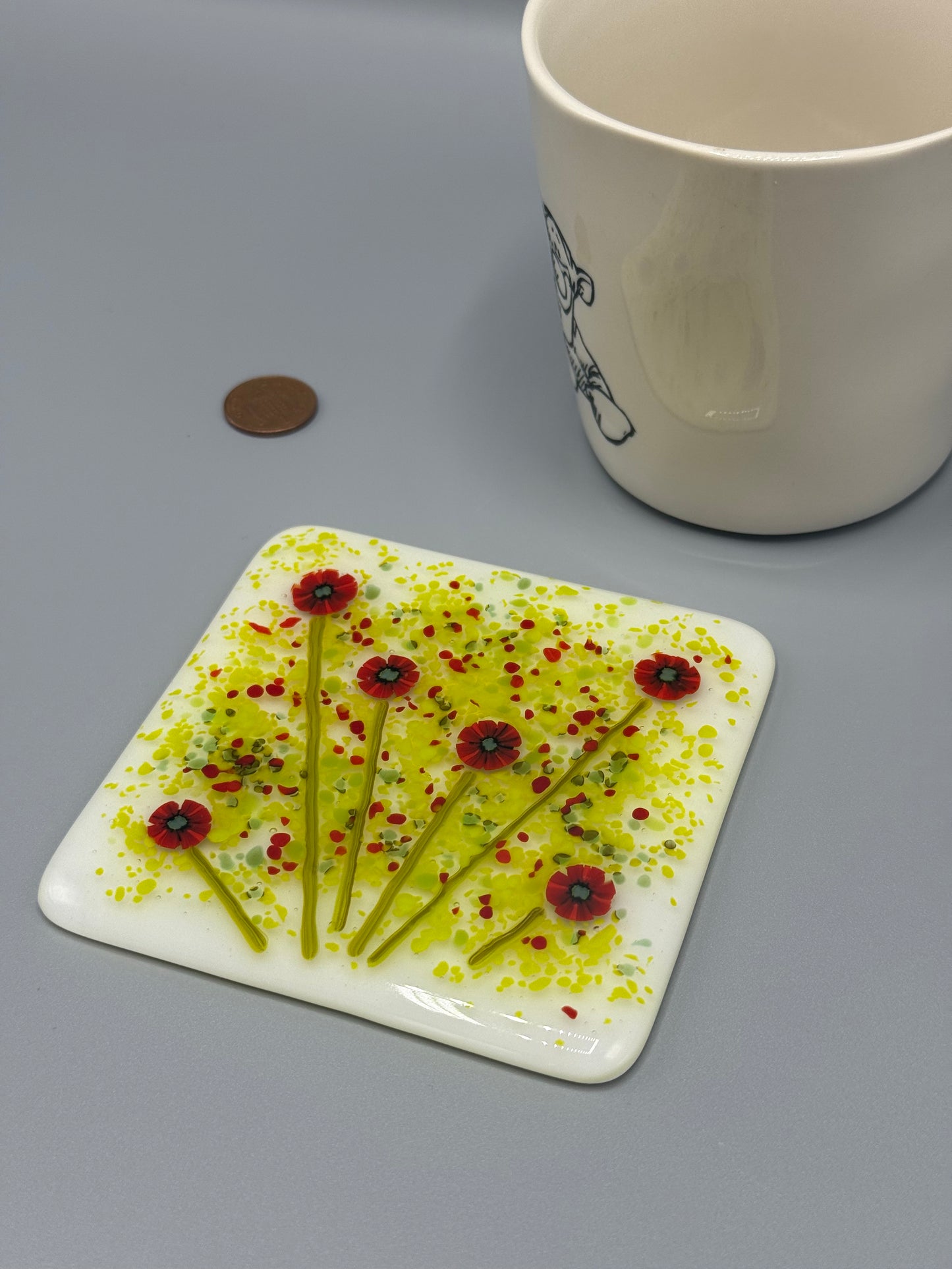 A square fused glass coaster with a poppy design, displayed next to a cup, with a coin on the side for scale.